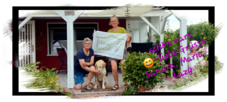 Zwei Frauen halten eine Fahne mit dem Logo des Ferienhauses und davor sitz ein Hund.